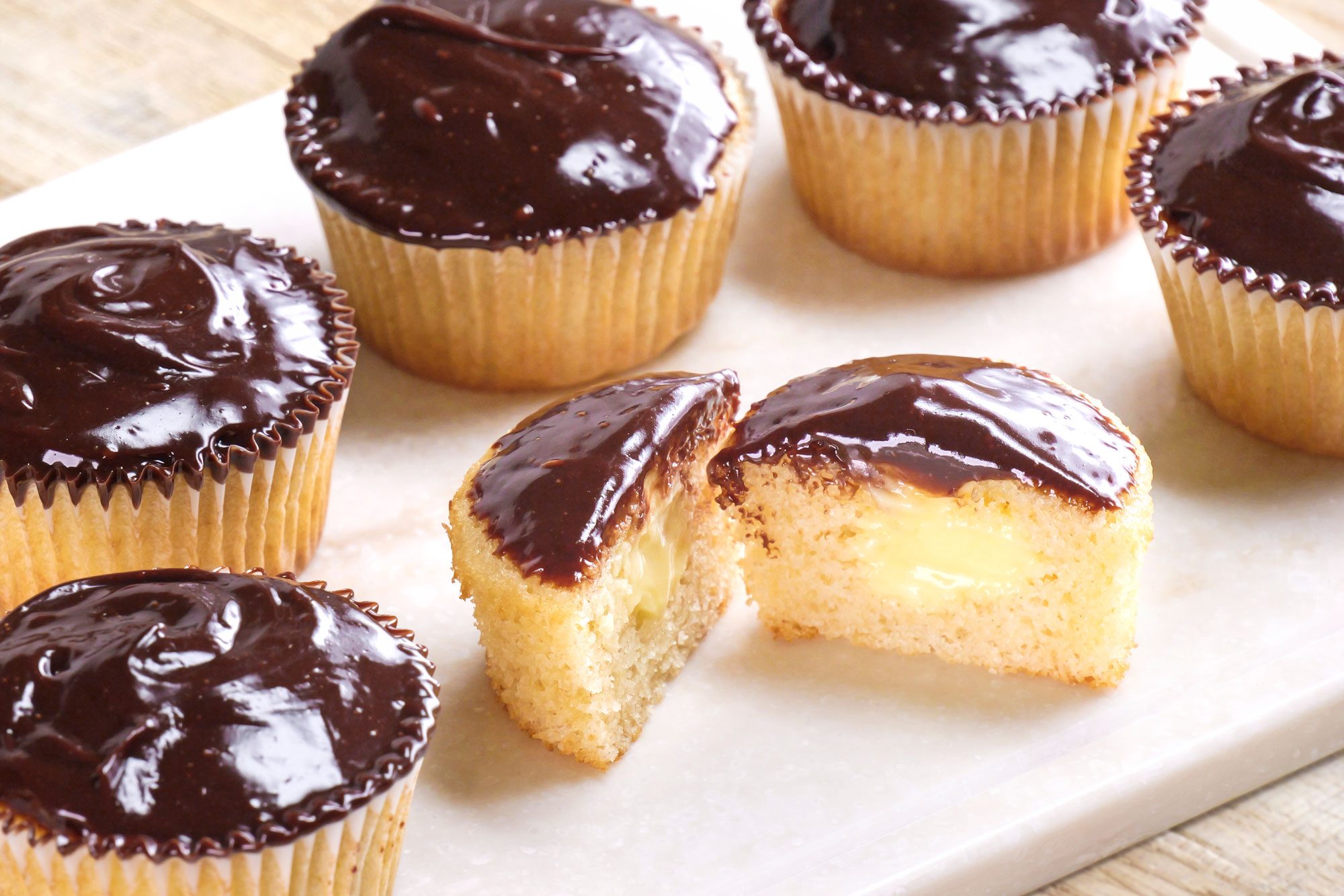 3/4th shot of Six Boston Cream Cupcakes with glossy chocolate frosting are arranged on a white surface, with one cupcake cut in half to reveal a creamy yellow filling inside, The background is a light wooden surface