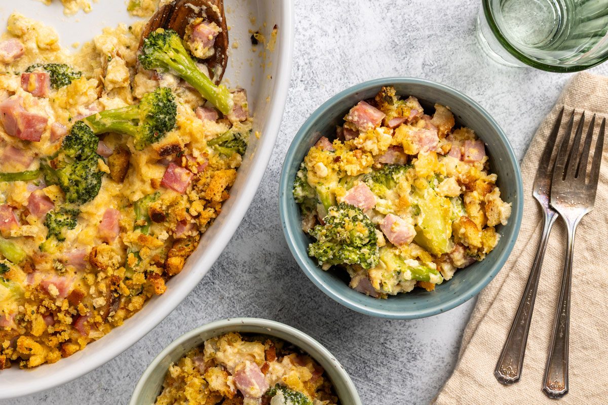 Top-down shot; a casserole dish with broccoli; diced ham; crumb topping beside two bowls of casserole; forks; glass of water on a light surface