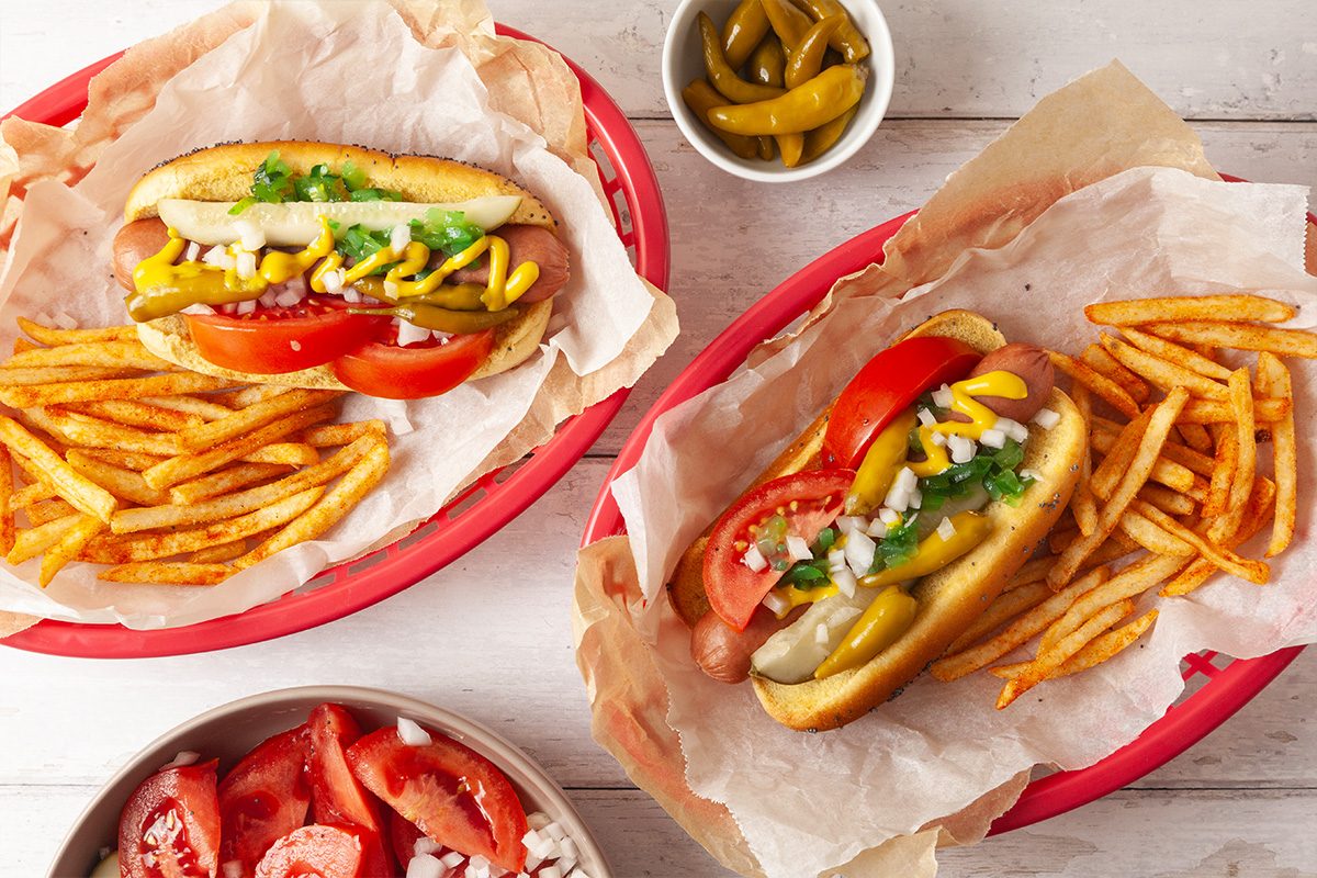 Two baskets with hot dogs topped with mustard, onions, pickles, tomatoes, and peppers, served with French fries. A small bowl of pickles and a bowl of chopped tomatoes and onions are nearby on a white wooden surface.