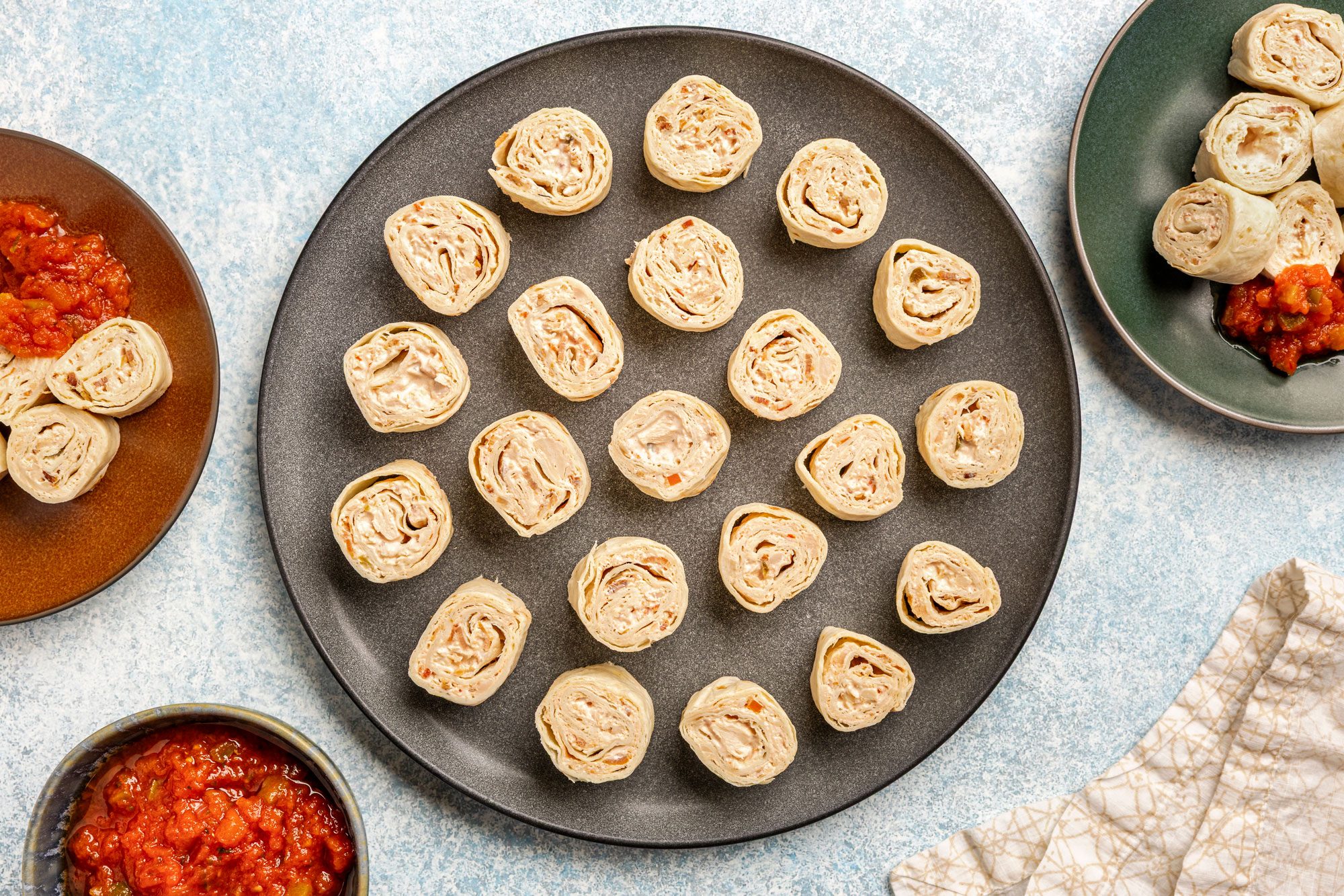 Overhead shot; a dark plate of pinwheel tortilla roll-ups on a light surface; nearby salsa bowls; plates; beige patterned napkin