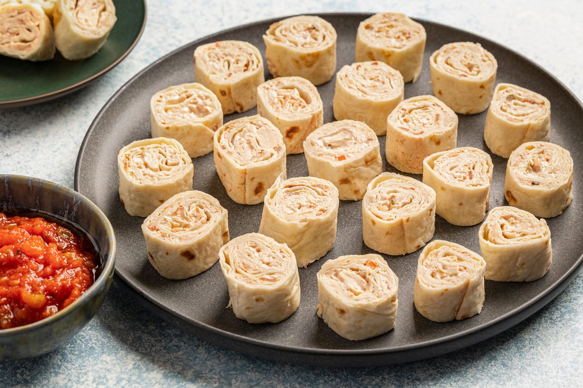 This is an overhead shot; a black plate has tortilla pinwheels with creamy filling; salsa and a smaller plate sit on a light blue surface