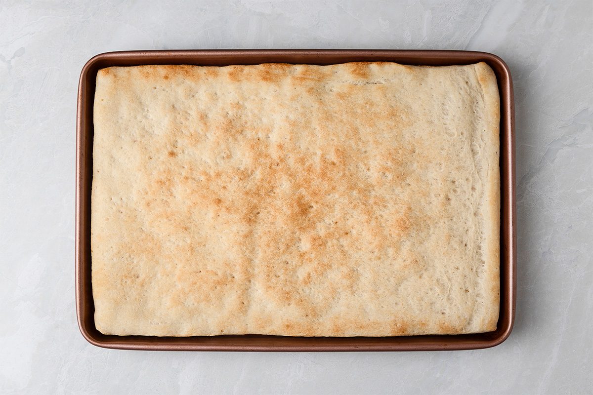 A rectangular sheet of golden-brown baked focaccia bread sits in a rimmed baking pan on a light gray surface.