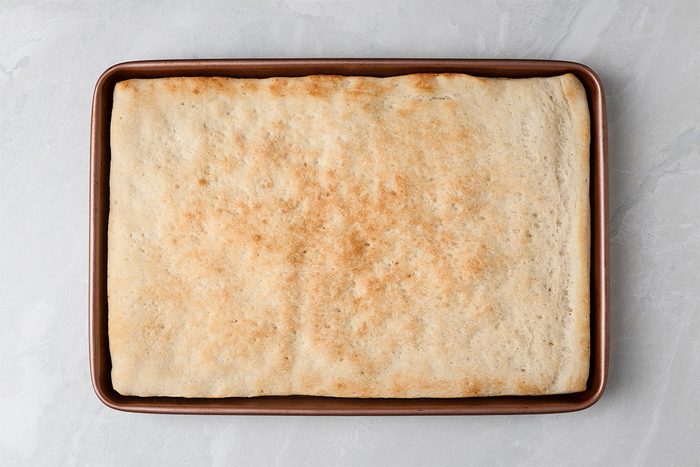 A rectangular sheet of golden-brown baked focaccia bread sits in a rimmed baking pan on a light gray surface.