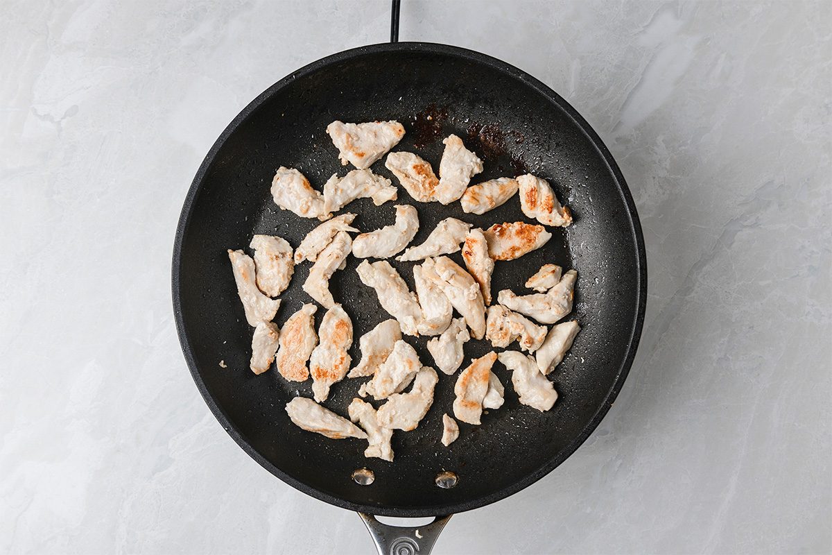 Strips of cooked chicken breast in a black frying pan on a light gray countertop, viewed from above.