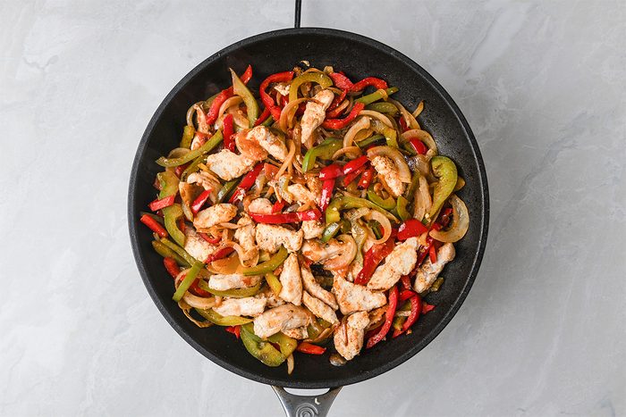 A skillet filled with cooked chicken strips, red and green bell peppers, and onions, all sautéed together and placed on a light gray countertop.