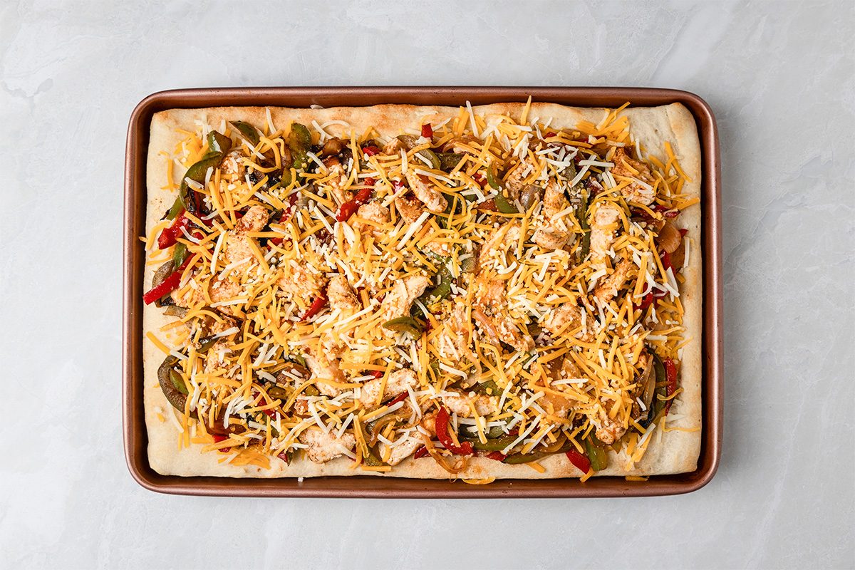 A rectangular baking sheet holds an unbaked pizza topped with shredded cheese, sliced peppers, chicken pieces, and other colorful ingredients on a light gray surface.