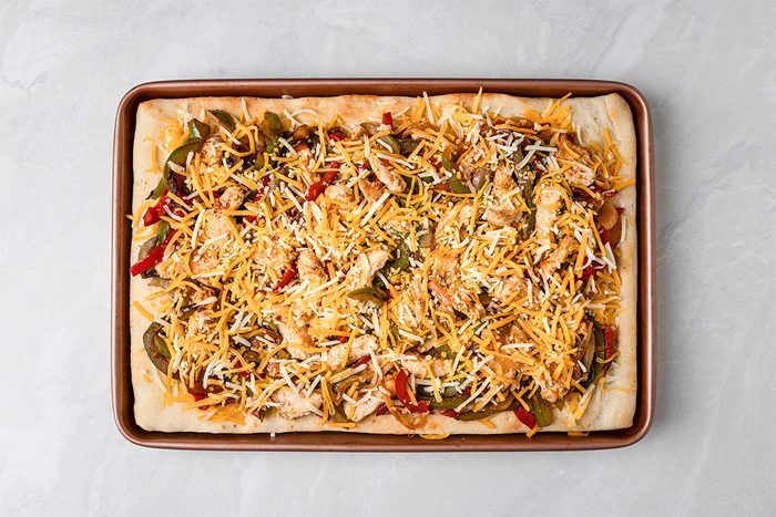 A rectangular baking sheet holds an unbaked pizza topped with shredded cheese, sliced peppers, chicken pieces, and other colorful ingredients on a light gray surface.