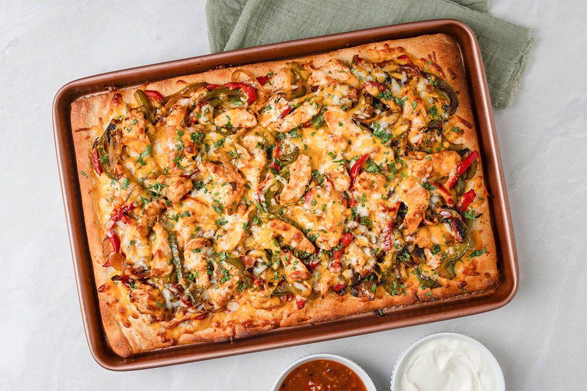 A rectangular pan pizza topped with melted cheese, sliced bell peppers, onions, and herbs sits on a baking tray. Nearby are small bowls of salsa and sour cream on a light gray surface.