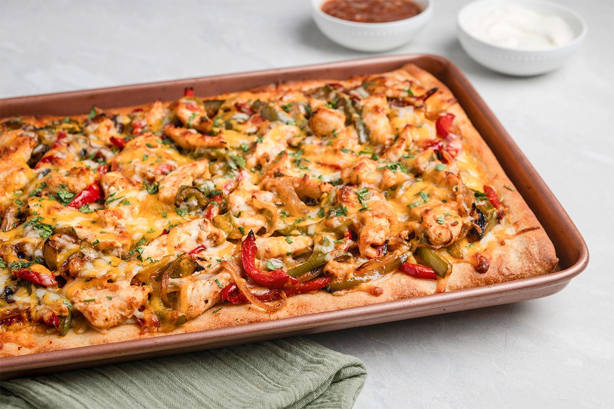 Rectangular sheet pan pizza topped with melted cheese, chicken pieces, red and green bell peppers, and herbs. In the background are two small bowls with sauces on a light surface.