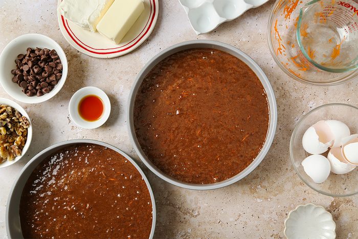 Two round cake pans filled with chocolate cake batter sit on a countertop surrounded by baking ingredients, including butter, cream cheese, eggs, chocolate chips, walnuts, vanilla extract, and a glass measuring cup.