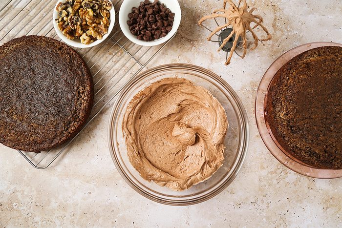 Two round chocolate cake layers, a bowl of chocolate frosting, bowls of walnuts and chocolate chips, and a plastic spider are arranged on a countertop for cake decorating.