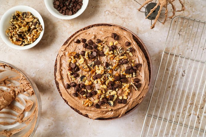 A chocolate cake topped with chocolate chips and chopped walnuts sits on a counter next to a bowl of walnuts, a bowl of chocolate chips, a mixing bowl with chocolate frosting, and a cooling rack.
