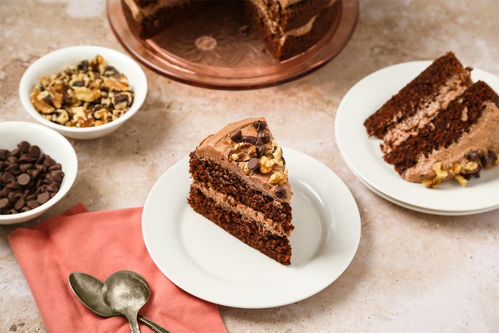 A slice of chocolate layer cake with chocolate frosting and walnuts sits on a white plate, next to a bowl of walnuts, a bowl of chocolate chips, two spoons, and a pink napkin. More cake is visible in the background.