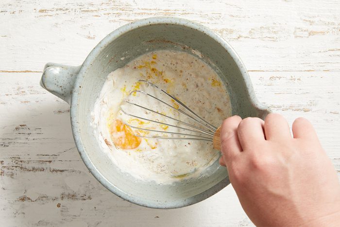 A hand whisking eggs, flour, and milk in a light gray mixing bowl on a white wooden surface.