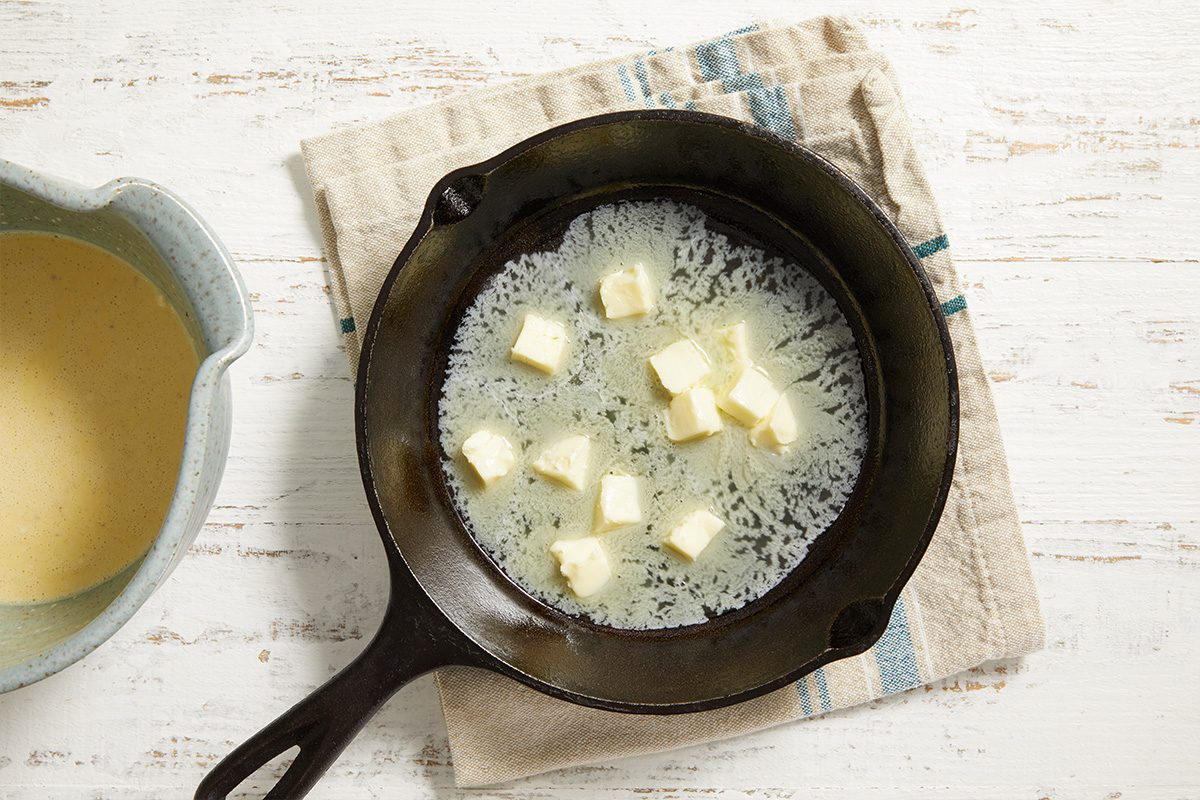 A cast iron skillet with several cubes of butter inside, sitting on a beige and blue striped cloth, next to a bowl of light yellow batter on a white wooden surface.