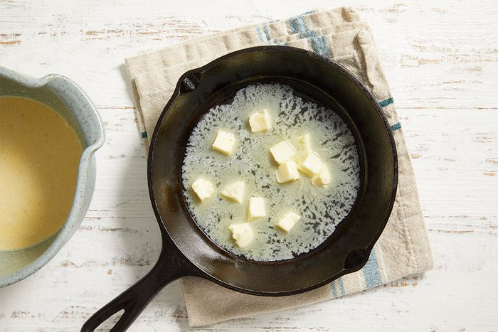 A cast iron skillet with several cubes of butter inside, sitting on a beige and blue striped cloth, next to a bowl of light yellow batter on a white wooden surface.