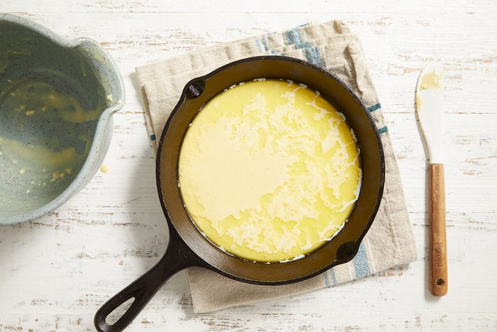 A cast iron skillet filled with cornbread batter sits on a folded kitchen towel. A mixing bowl and a butter knife are nearby on a white wooden surface.