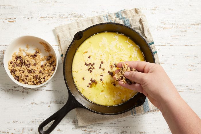 A hand sprinkles a crumbly topping over a yellow batter in a black cast-iron skillet, with a bowl of topping mix nearby on a white wooden surface.