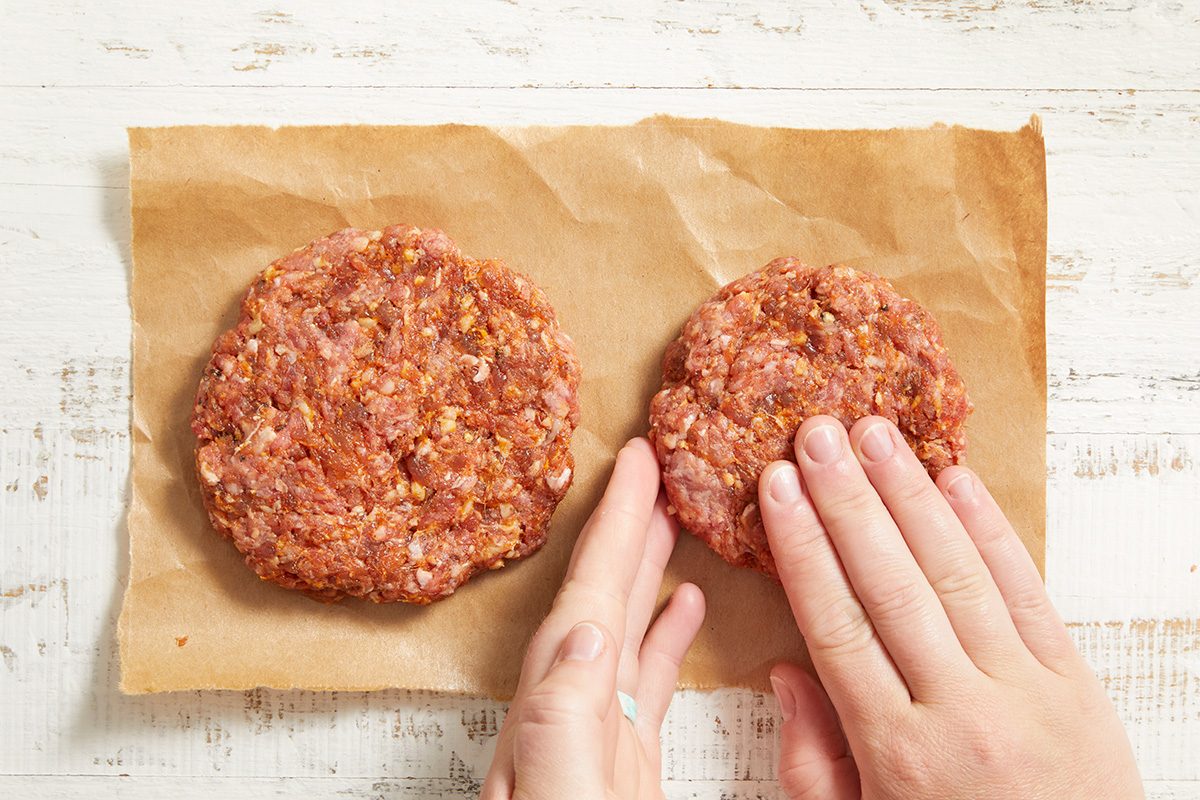 Two hands shaping raw ground meat into burger patties on a sheet of brown parchment paper, placed on a white wooden surface.