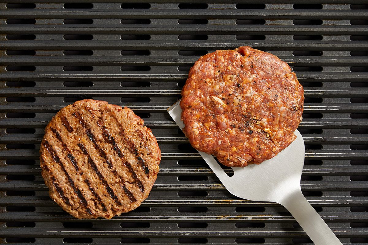 A raw hamburger patty on a spatula sits next to a cooked patty with grill marks on a barbecue grill.