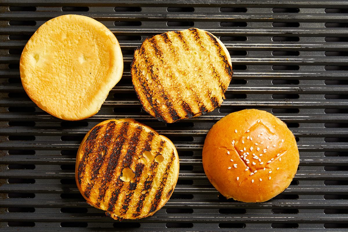 Four hamburger bun halves on a grill, two with visible dark grill marks, one plain top bun with sesame seeds, and one plain bun bottom without grill marks.
