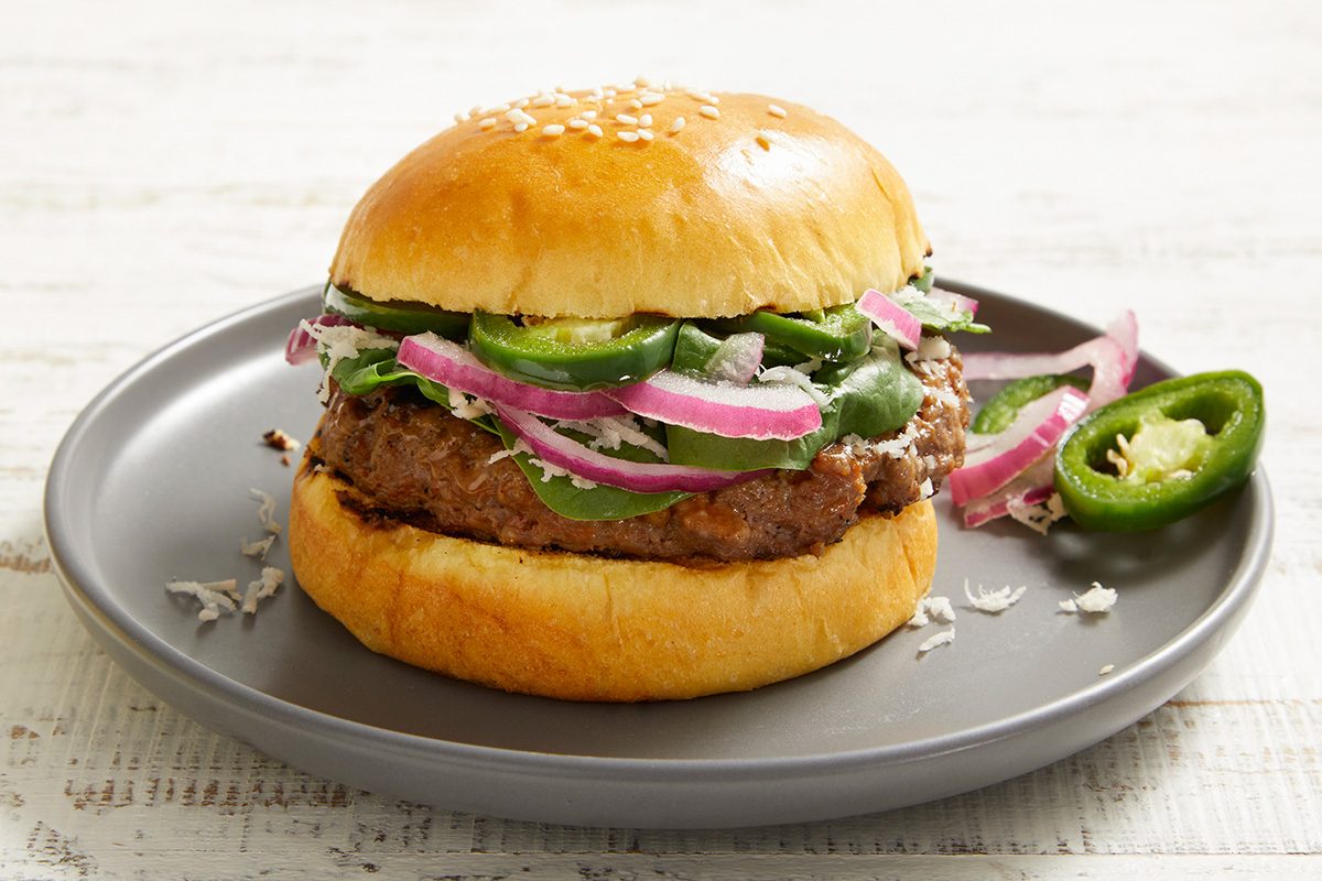 A hamburger on a gray plate, topped with sliced red onions, jalapeños, lettuce, and cheese in a sesame seed bun, with extra jalapeño slices on the side.