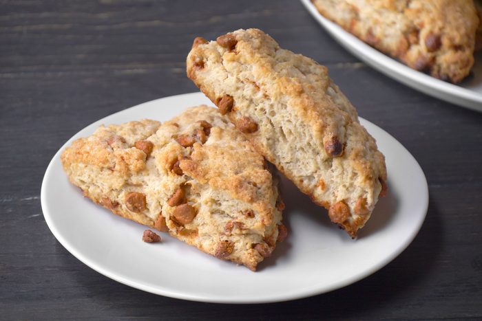 3/4th shot of Two golden brown scones with visible cinnamon chips sit on a white plate, with more scones on a plate in the background, set on a dark wooden surface