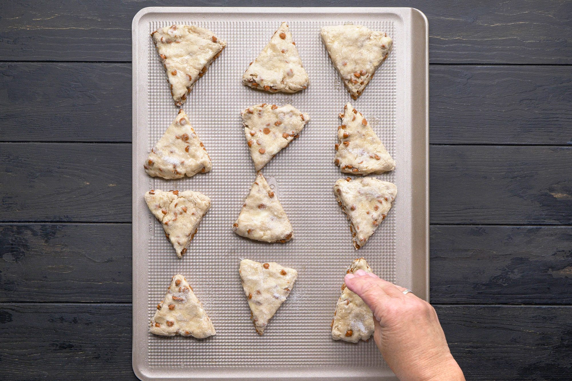 overhead shot of A hand places a triangle shaped, unbaked scone onto a baking sheet lined with parchment; Eleven other scones are already arranged on the sheet, ready for baking, atop a dark wooden surface
