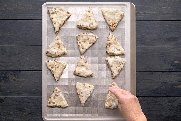 overhead shot of A hand places a triangle shaped, unbaked scone onto a baking sheet lined with parchment; Eleven other scones are already arranged on the sheet, ready for baking, atop a dark wooden surface