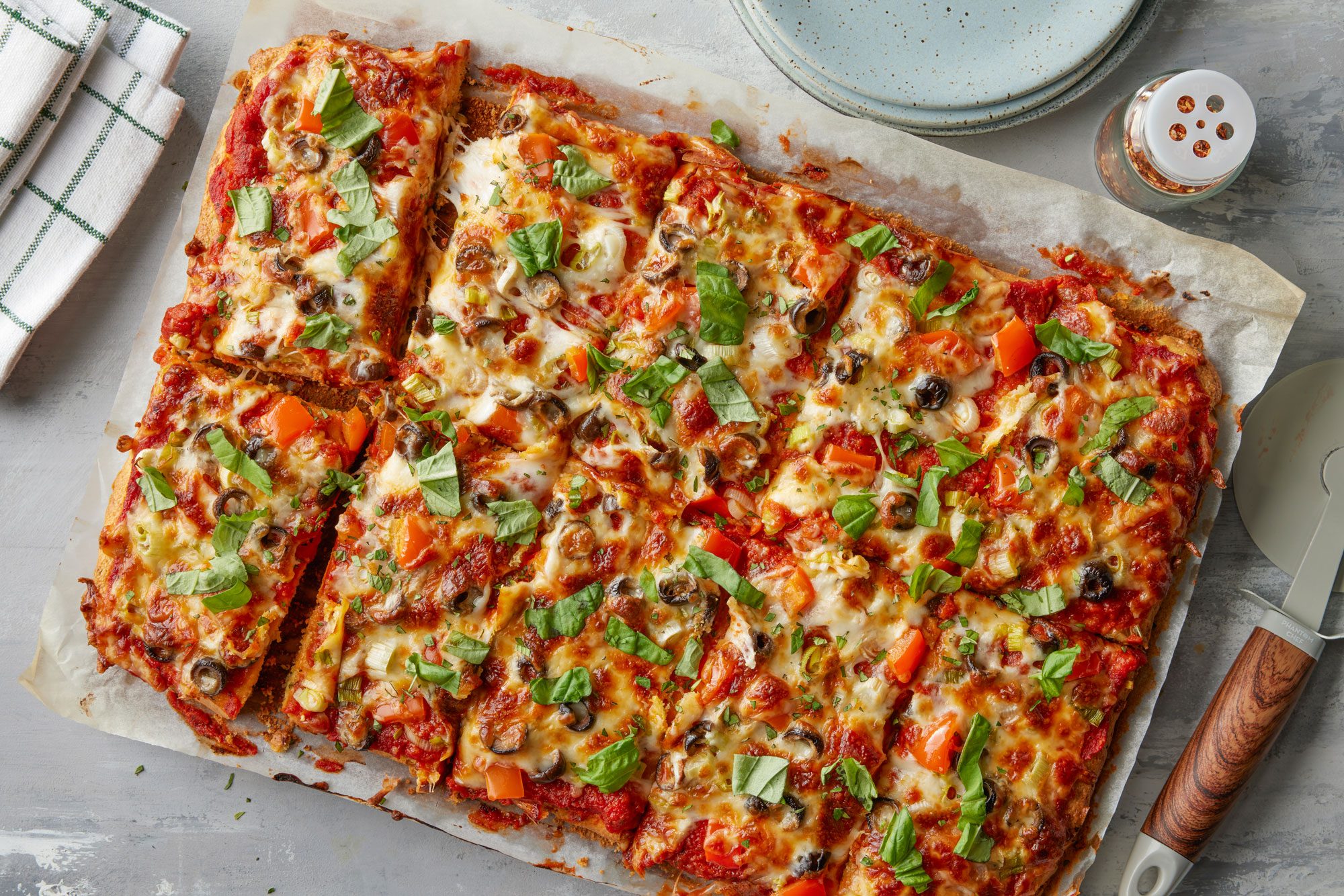 overhead shot of Rectangular sheet pan pizza topped with melted cheese, chopped basil, olives, red bell peppers, and tomato sauce sits on parchment paper; A pizza cutter, spice jar, and stack of plates are nearby
