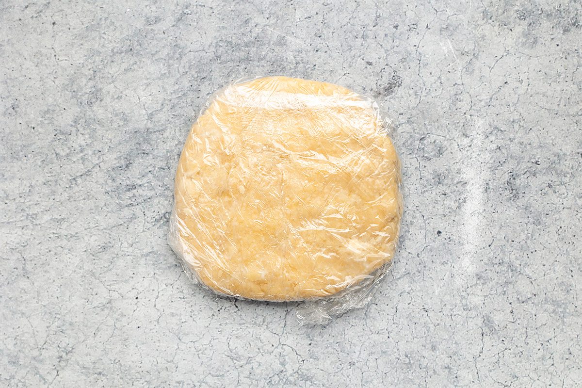 A disk of pale yellow dough wrapped tightly in plastic wrap, resting on a light gray, textured surface.