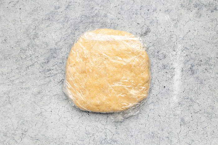 A disk of pale yellow dough wrapped tightly in plastic wrap, resting on a light gray, textured surface.