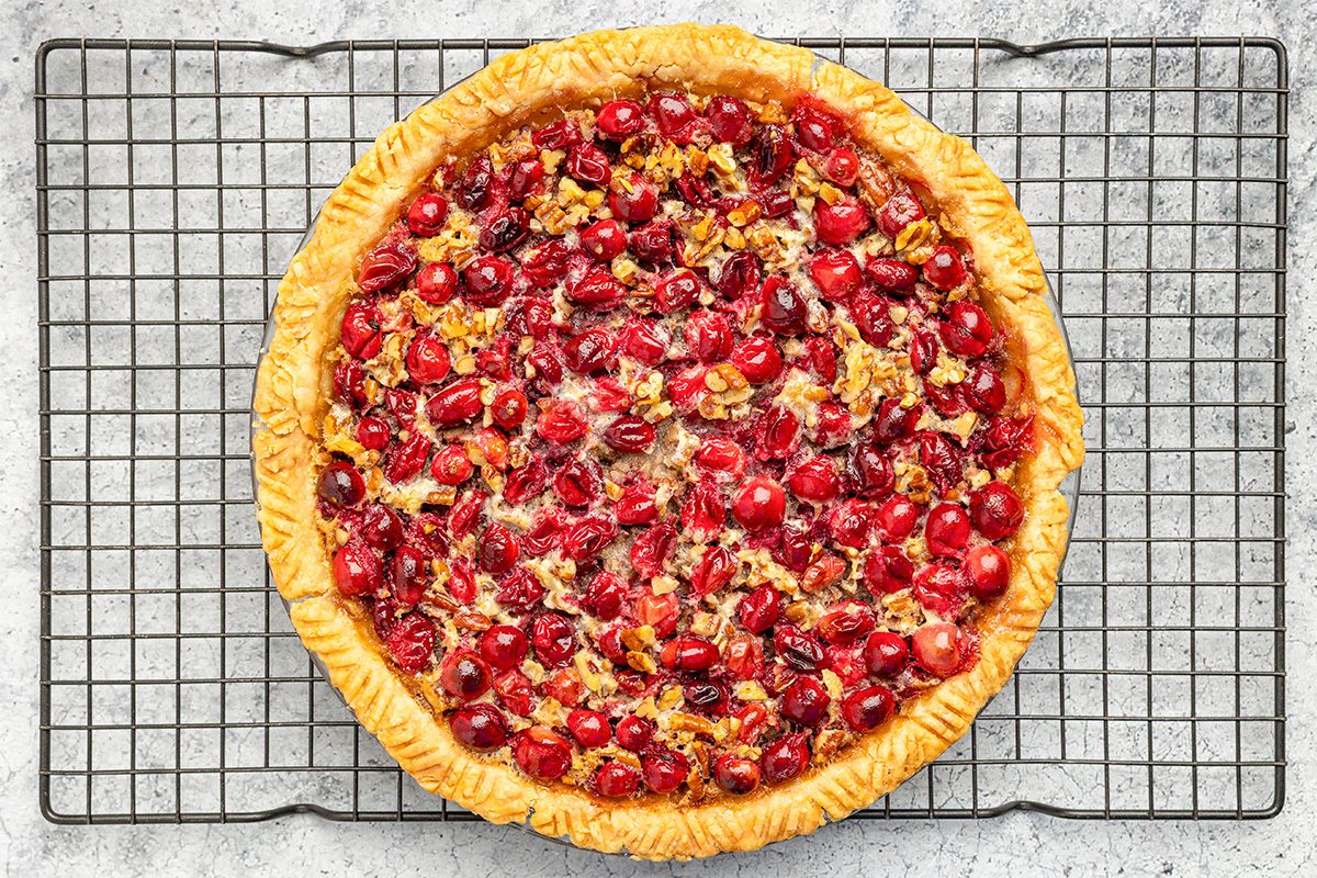 A freshly baked cranberry pie with a golden crust sits on a wire cooling rack, filled with vibrant red cranberries and chopped nuts on a light gray surface.
