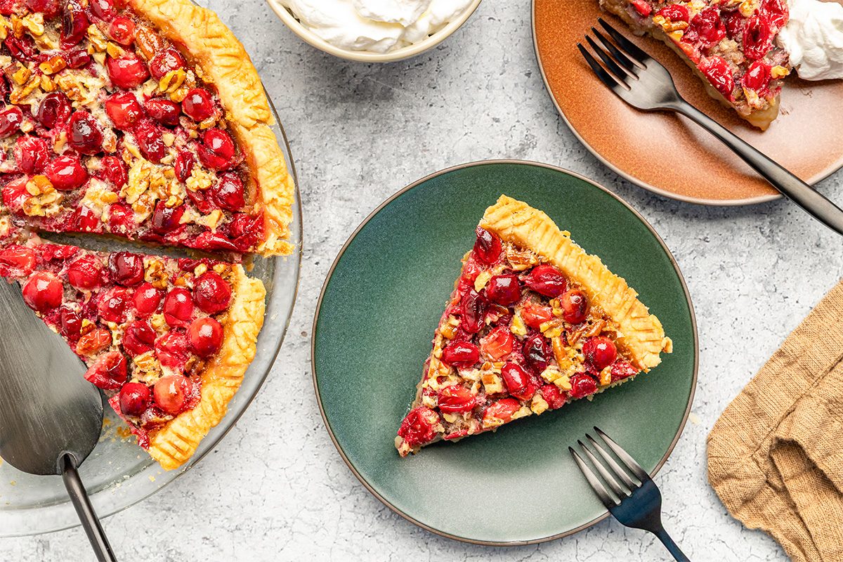 A cherry pecan pie with a golden crust sits on a glass plate, with two slices served on green and orange plates. A bowl of whipped cream and utensils are nearby on a light stone surface.