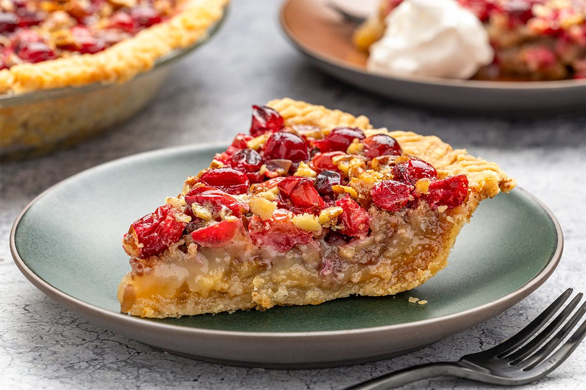 A slice of fruit pie with a golden crust, filled with red cranberries and chopped nuts, sits on a green plate. A fork is nearby and another slice with whipped cream is in the background.