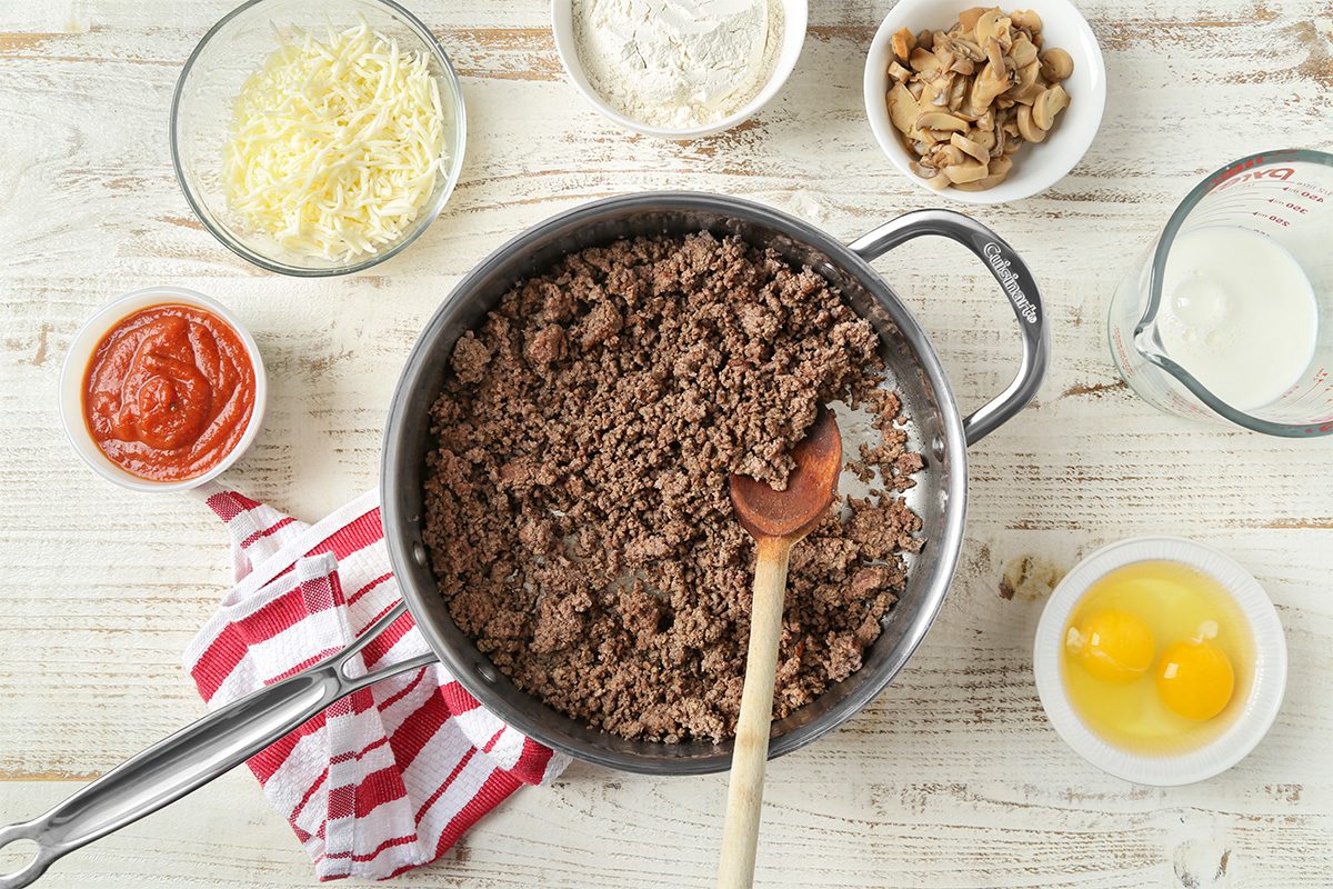 Overhead view of a pan with cooked ground beef and a wooden spoon, surrounded by bowls of shredded cheese, tomato sauce, flour, mushrooms, milk, and two cracked eggs on a rustic white surface.
