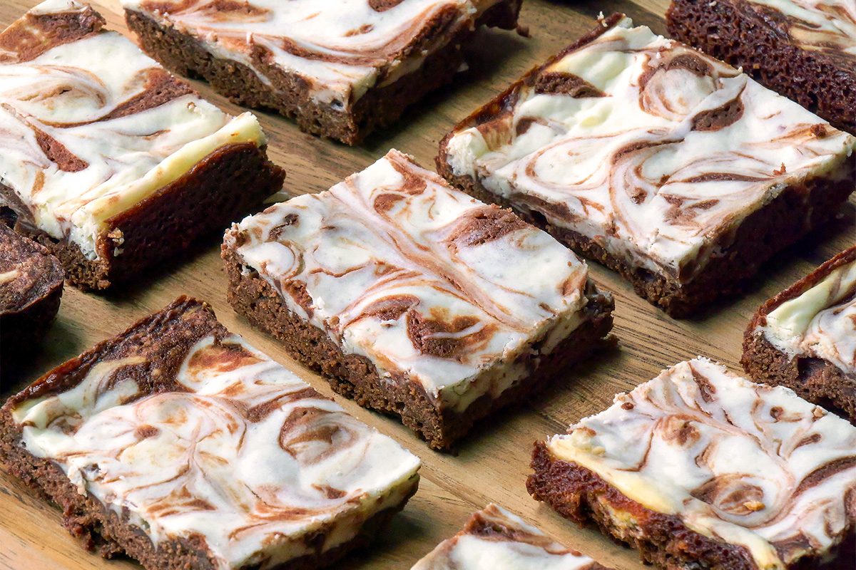 A close-up of several cheesecake brownies arranged on a wooden surface. Each brownie has a marbled swirl of creamy white cheesecake and rich chocolate brownie layers.