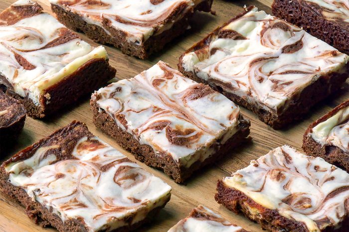 A close-up of several cheesecake brownies arranged on a wooden surface. Each brownie has a marbled swirl of creamy white cheesecake and rich chocolate brownie layers.