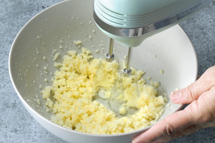 A hand holds a white mixing bowl while using a mint green electric hand mixer to cream together butter and sugar; the mixture appears pale and grainy.