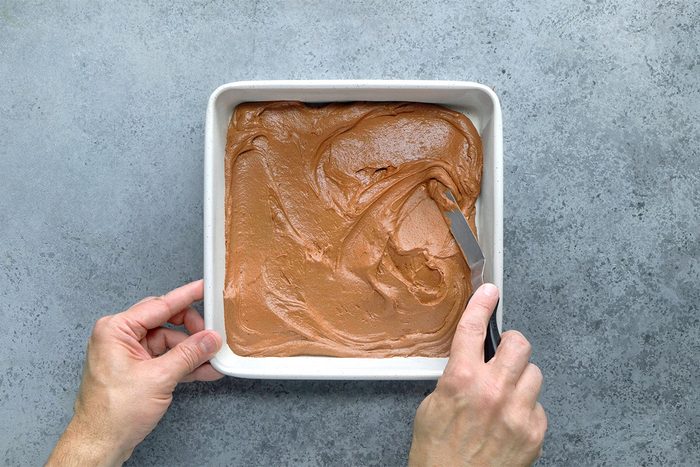 A person spreads chocolate cake batter evenly in a square baking pan with a spatula, on a gray countertop.