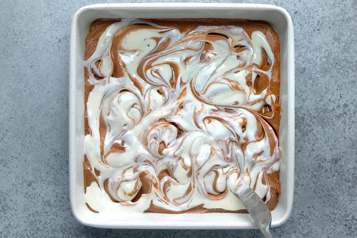 A square baking dish filled with swirled chocolate and white batter, with a spatula resting in one corner, on a gray surface.