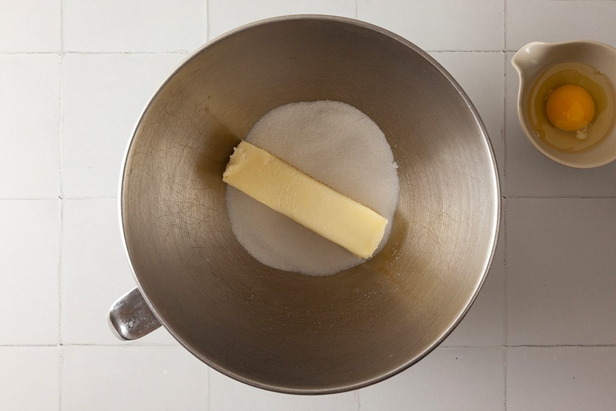A stick of butter and granulated sugar are in a metal mixing bowl, with a small bowl holding a cracked egg beside it on a white tiled surface.