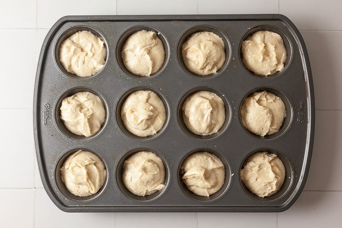 A muffin tin filled with twelve portions of unbaked muffin batter, ready to be baked, sits on a white tiled surface.