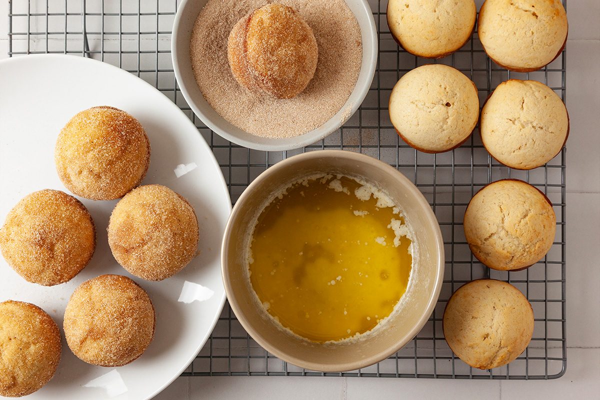 Baked muffins on a cooling rack, some coated with cinnamon sugar, with a bowl of melted butter and another bowl of cinnamon sugar nearby. A plate holds finished muffins.