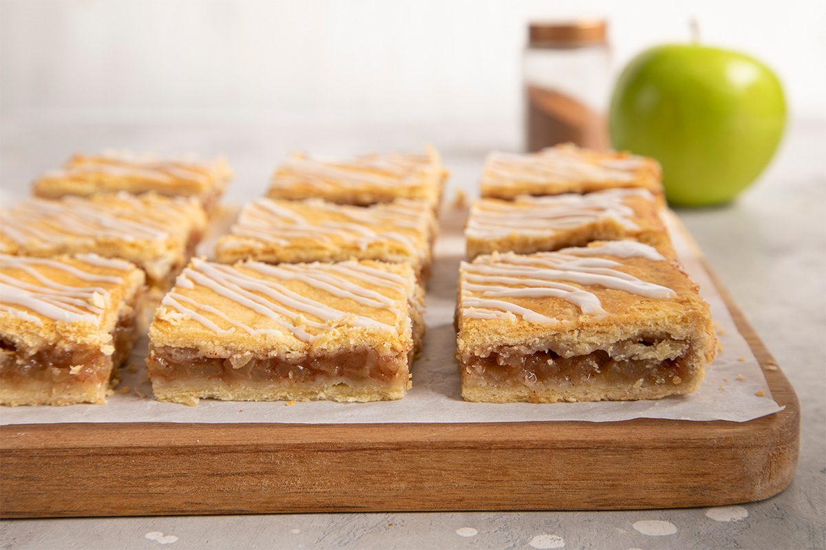 Rectangular apple bars with a golden crust and drizzled white icing are arranged on a wooden board. In the background, there is a green apple and a jar of spice.