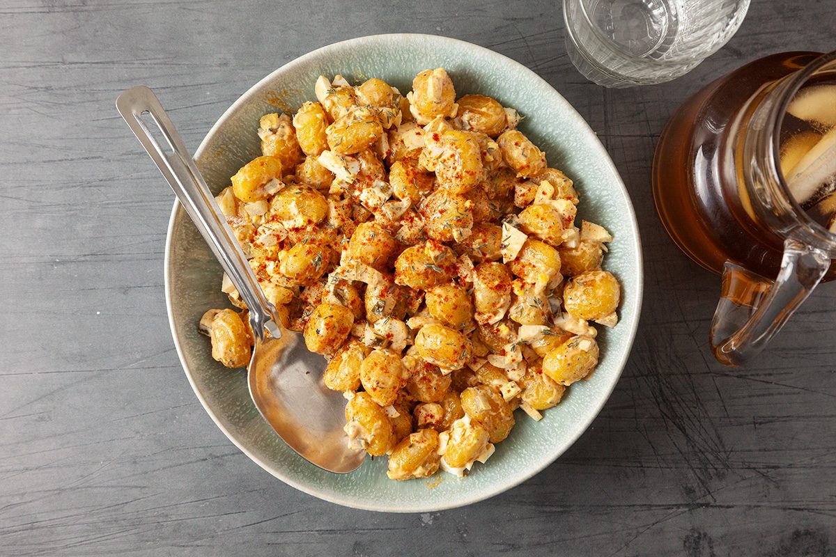 A bowl of cooked gnocchi topped with crumbled cheese and spices, with a spoon resting inside. Next to the bowl are a glass of iced drink and a pitcher on a gray tabletop.