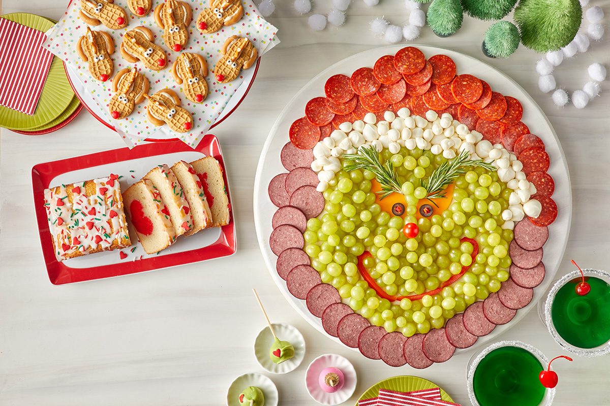 Overhead shot of a festive table features a large platter arranged to resemble the Grinch's face with grapes, mozzarella balls, and meats, surrounded by Christmas themed cookies, snack cake slices, and two green drinks with cherries; all set on a white table