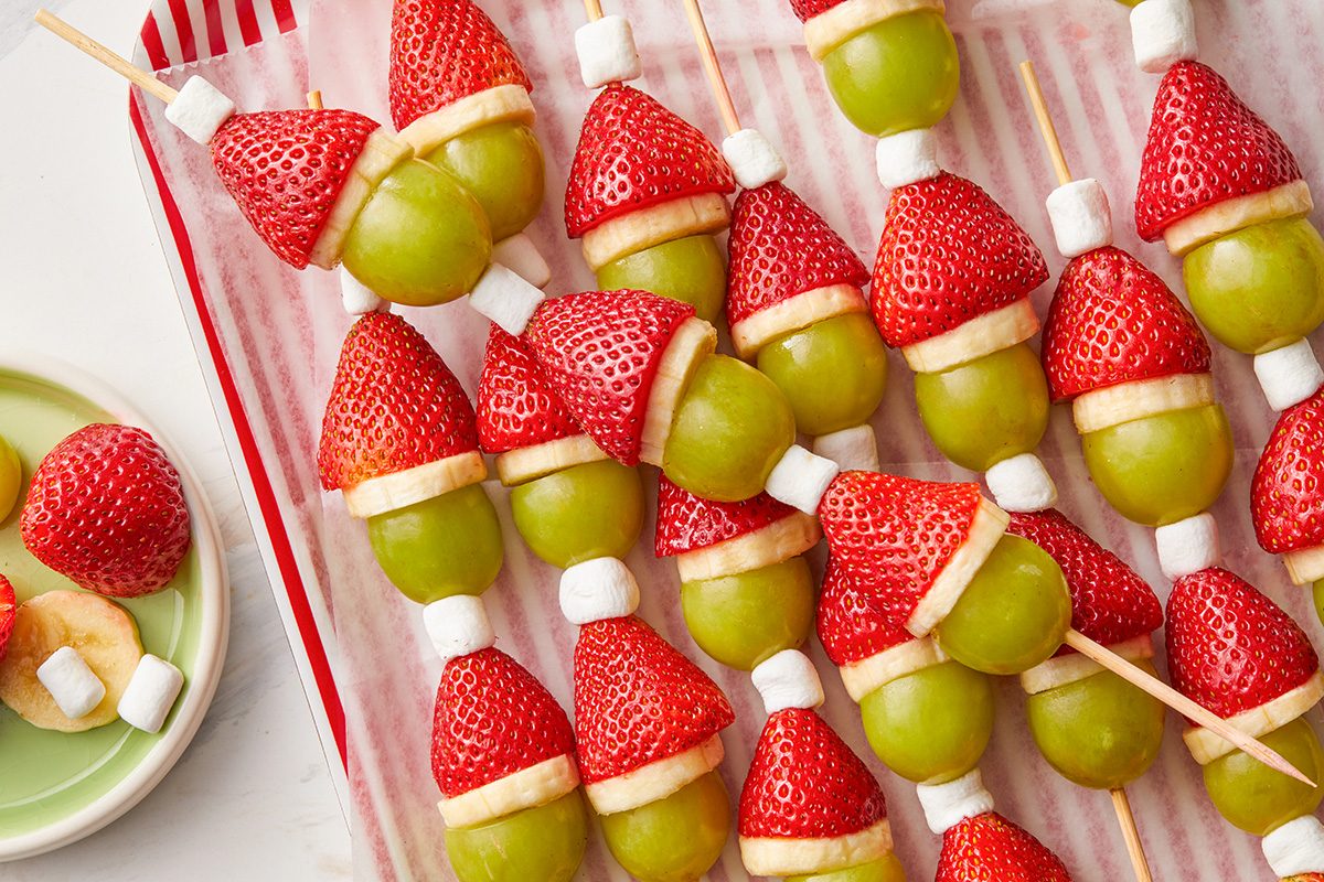overhead shot of Skewers with fruit arranged to look like Santa hats, using strawberries, green grapes, banana slices, and mini marshmallows, displayed on a striped tray