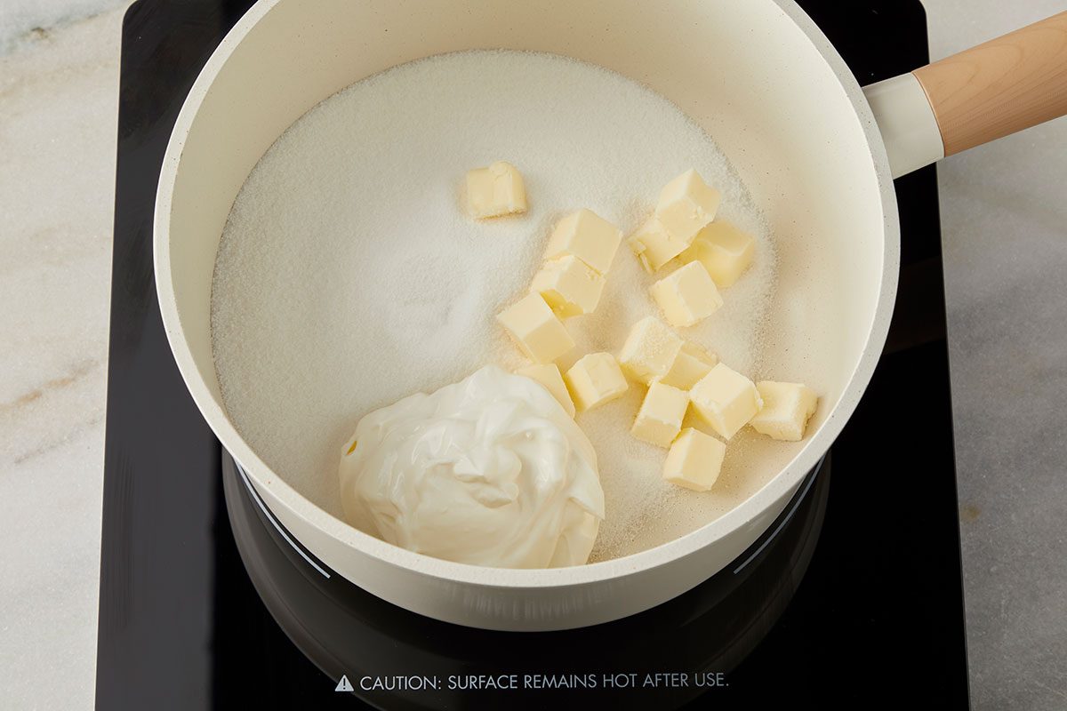 3/4th shot of a cream colored saucepan on a stovetop contains granulated sugar; cubed butter and a dollop of sour cream