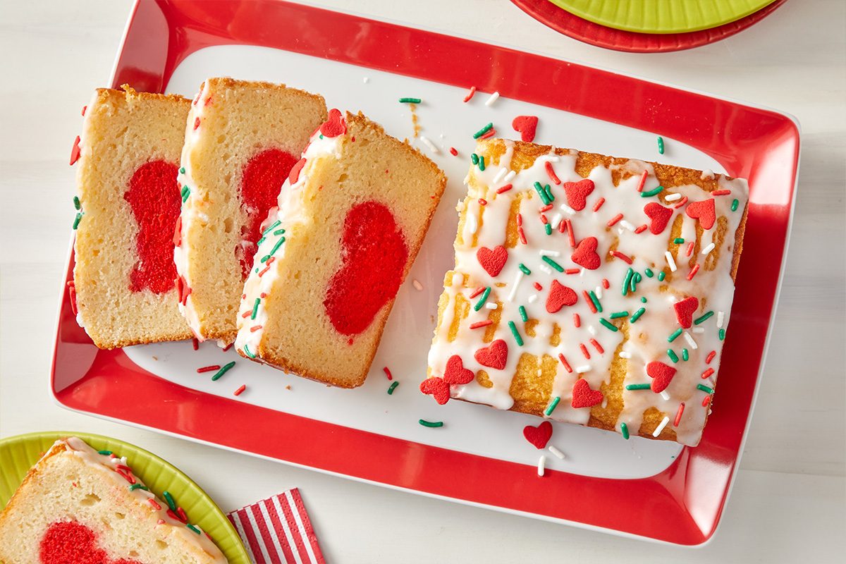 A rectangular loaf cake with white icing and red and green sprinkles sits on a red-edged platter. Three slices reveal a red heart shape inside each piece. Nearby are colorful plates and a striped napkin.